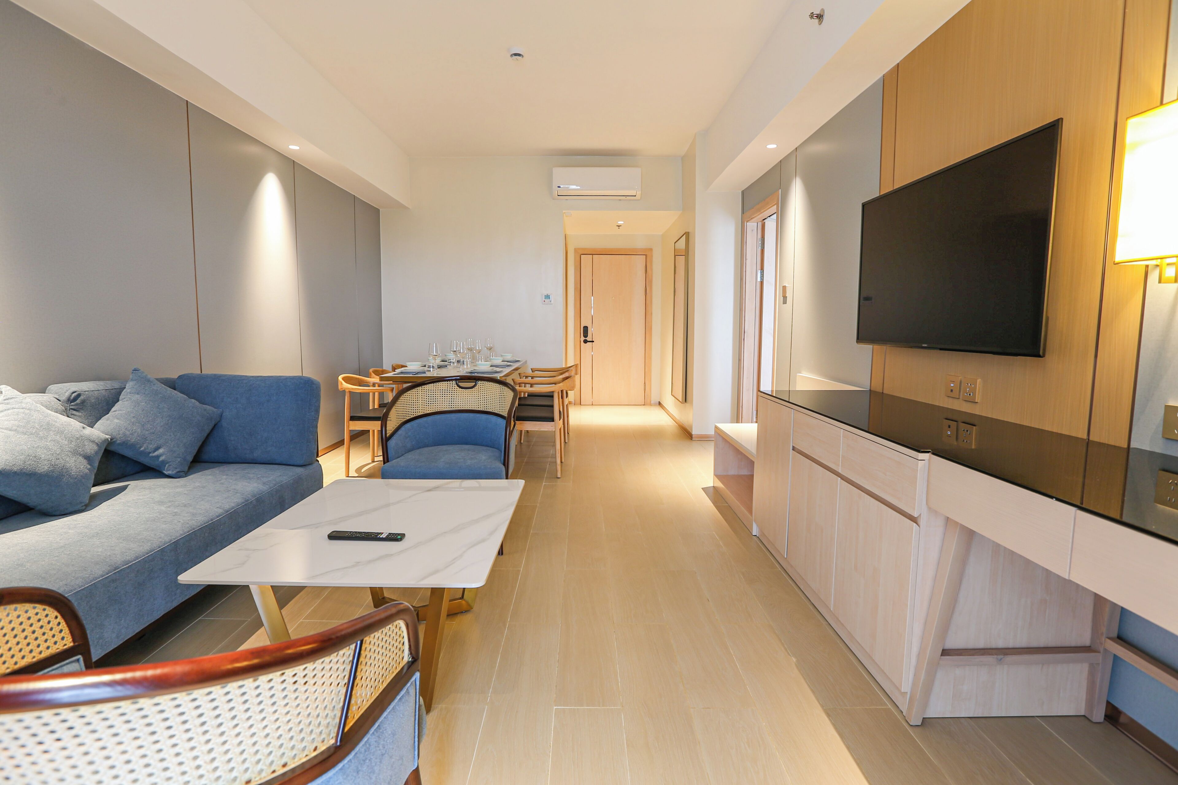 A modern living room with a sofa, a dining table with chairs, a kitchen area with a TV, and a hallway leading to a door.