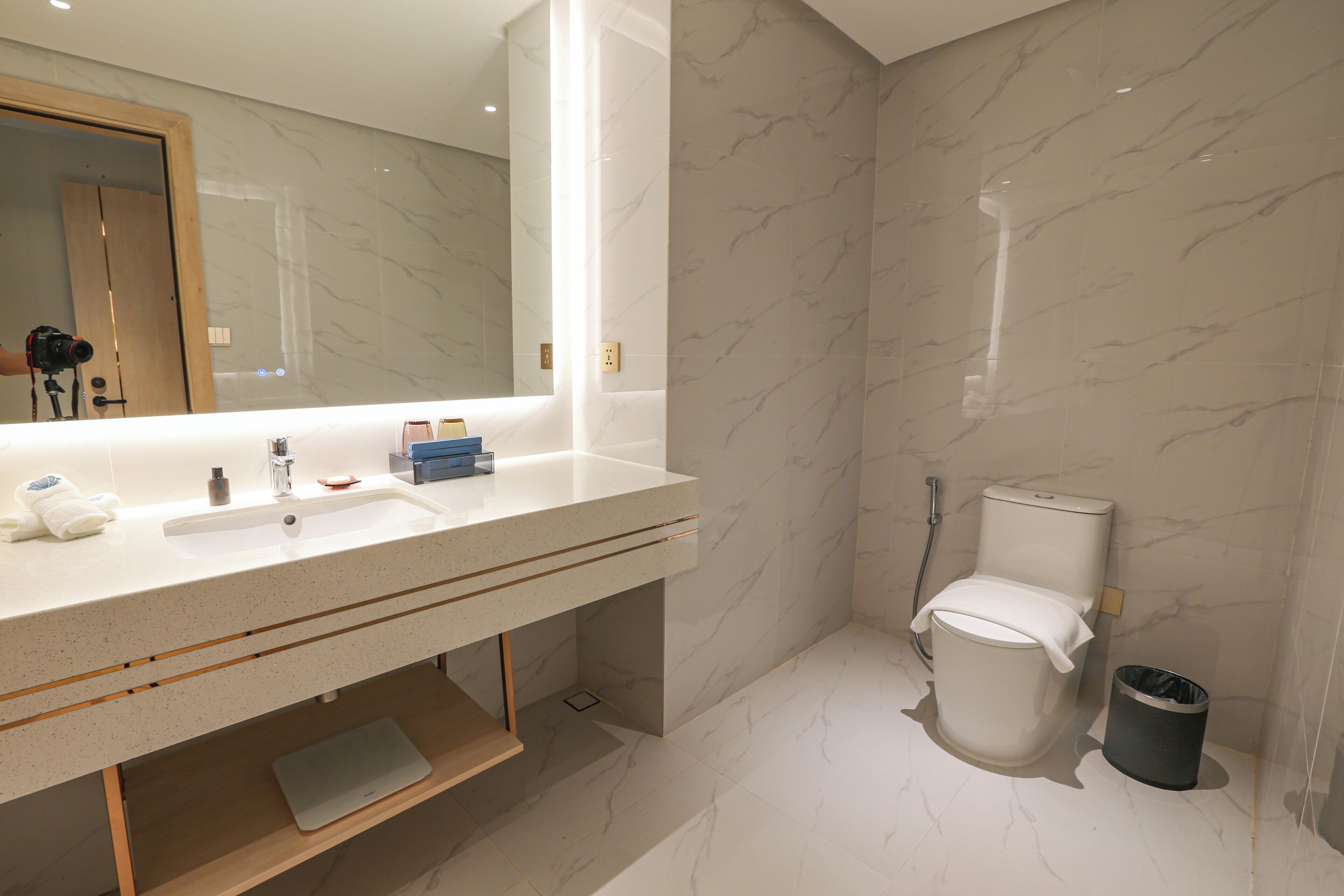 A modern bathroom with a marble sink, a toilet, a shower, and a trash bin.