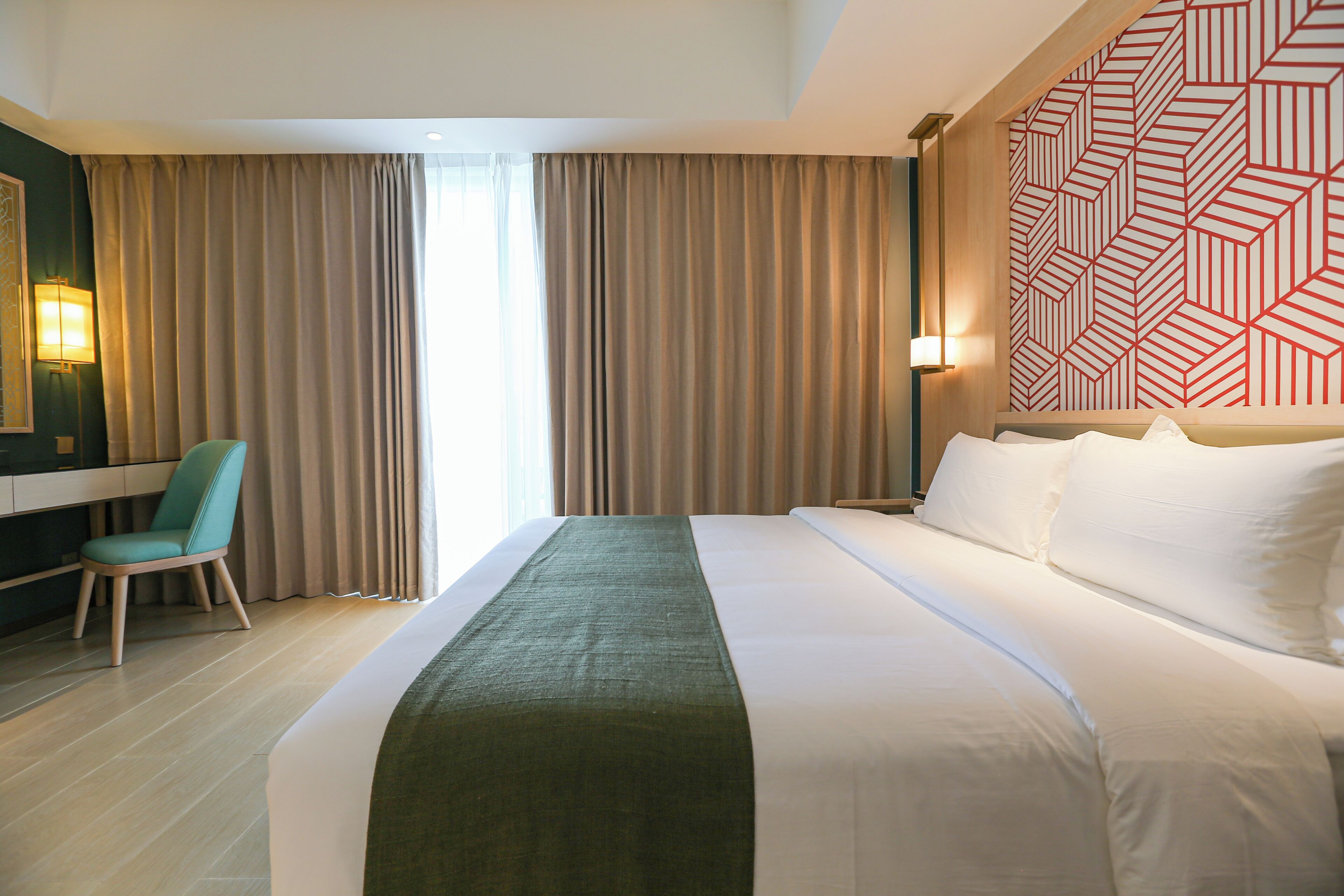 Una habitación de hotel con cama, escritorio, silla y una ventana grande con cortinas.