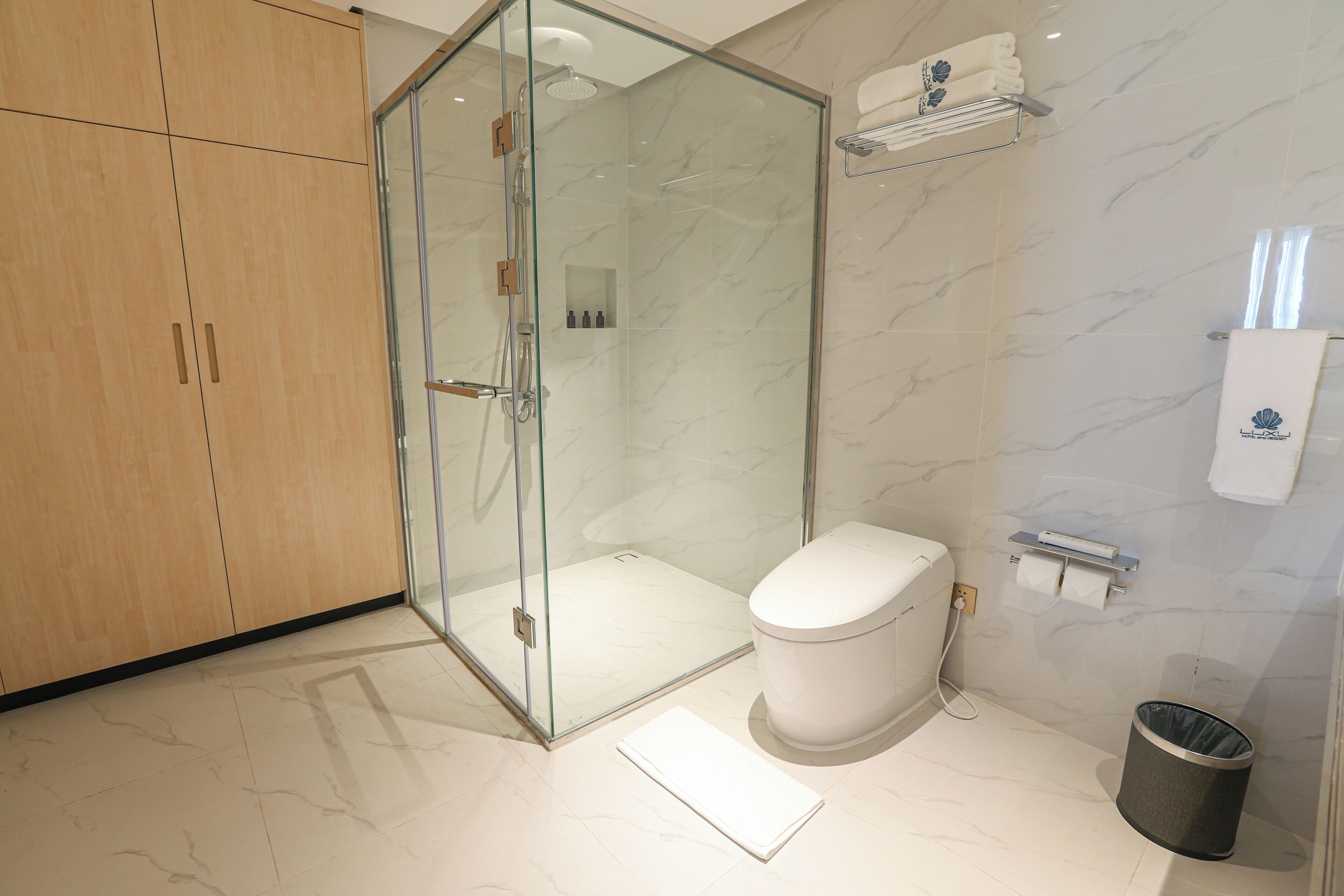 A modern bathroom with a glass-enclosed shower, a toilet, a trash can, and a towel rack.