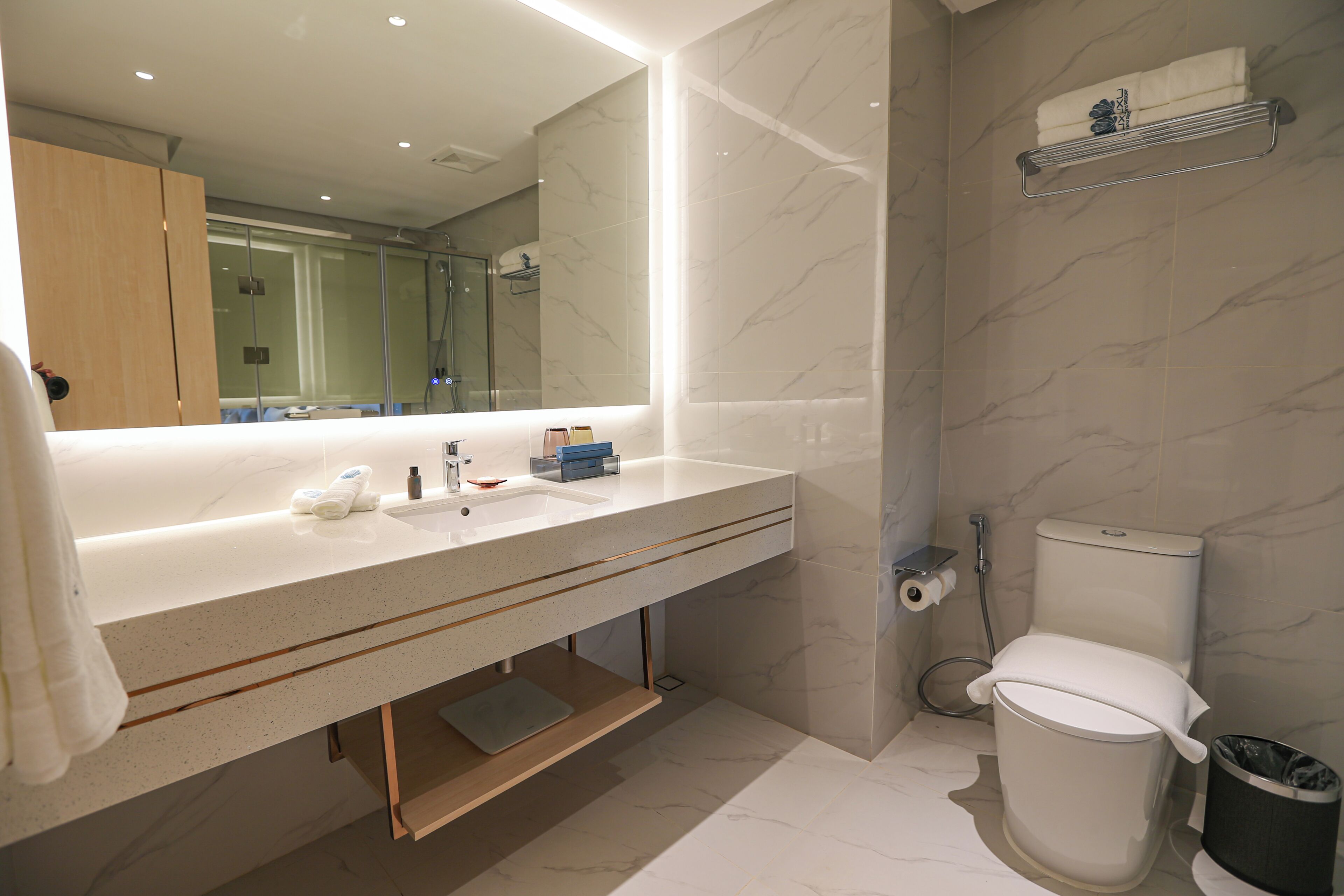A modern bathroom with a marble sink, a large mirror, a toilet, and a trash bin.