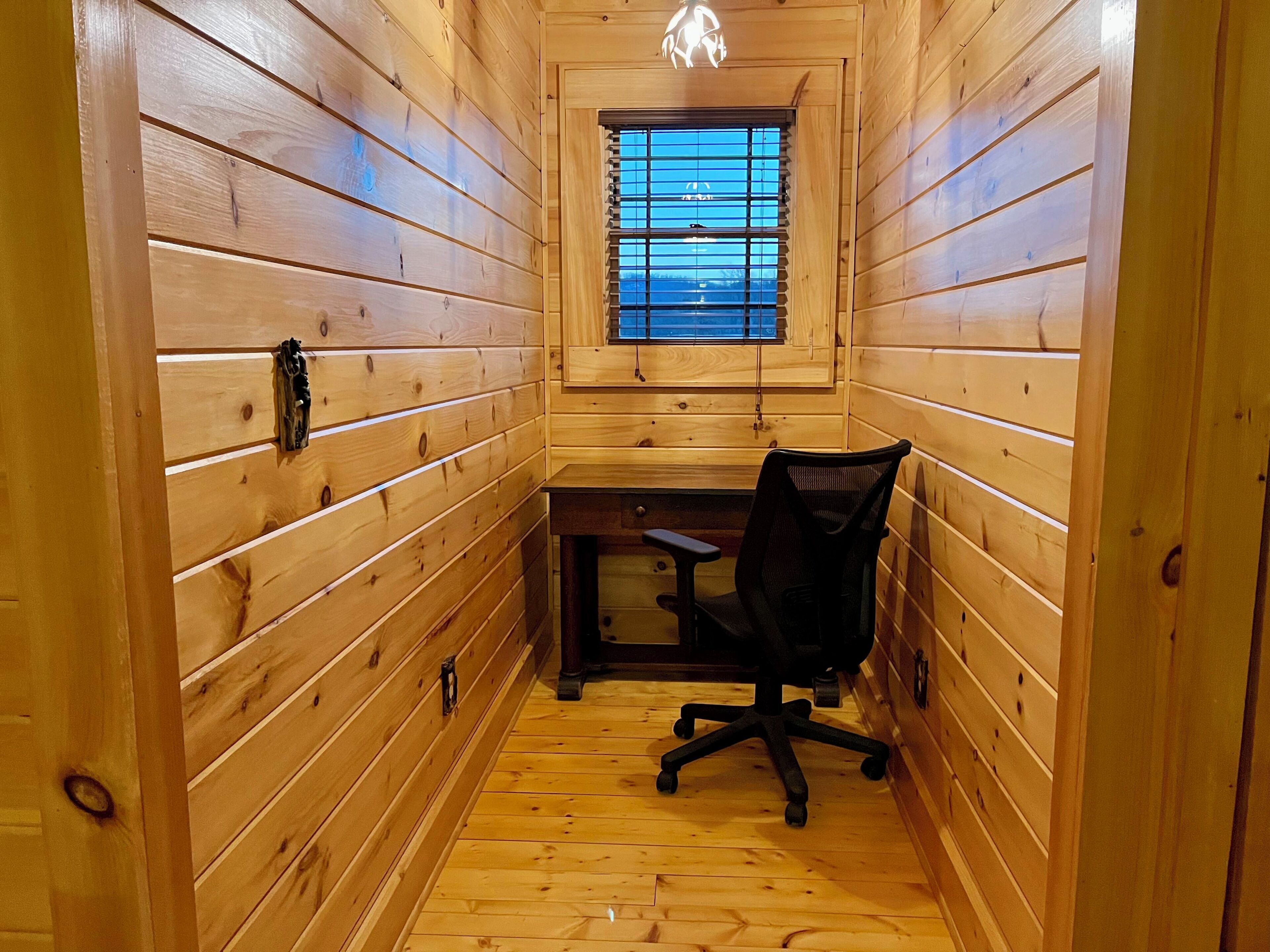 A narrow, wooden-paneled room with a desk and office chair, a window with blinds, and a hanging light fixture.