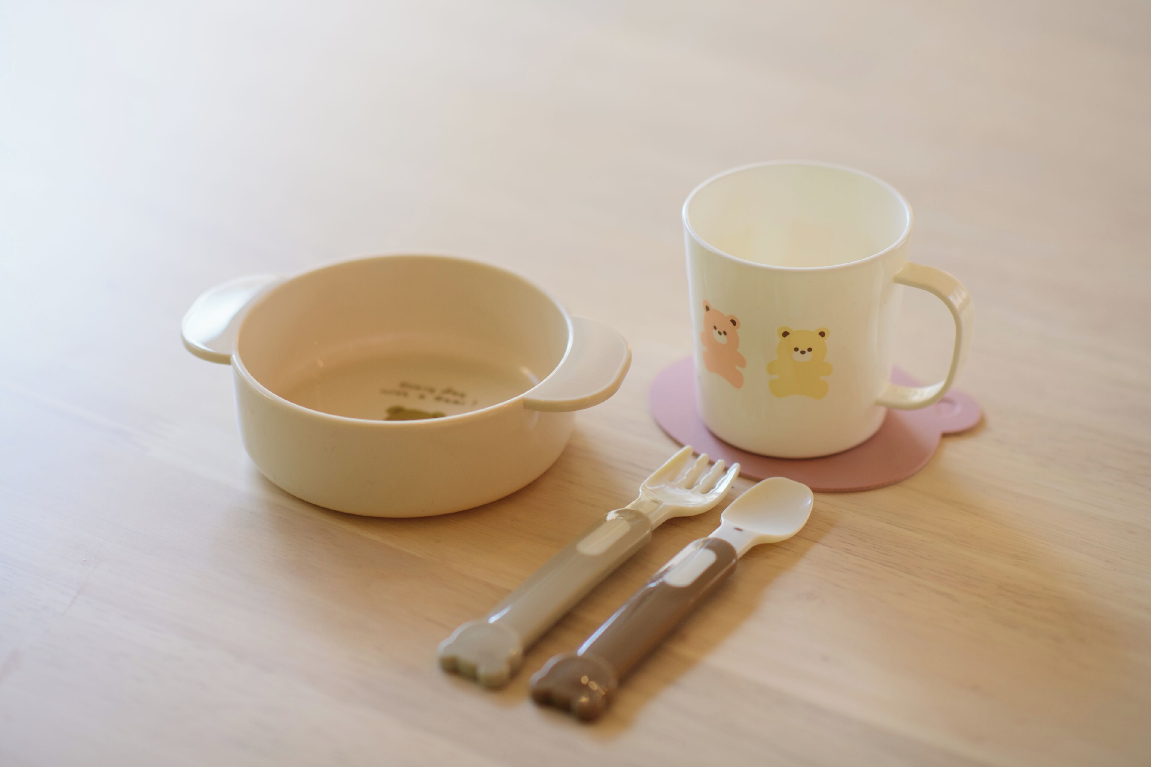 Eine beigefarbene Schale mit Deckel, eine Tasse mit Bär-Motiv sowie ein Gabel- und Löffelset auf einem Holztisch.