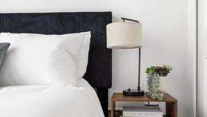 A neatly made bed with white bedding, a black headboard, a bedside lamp, and a small wooden nightstand with books and a plant. - Modern Loft in Vibrant Downtown Royal Oak - Just a Block Away! (Royal Oak)