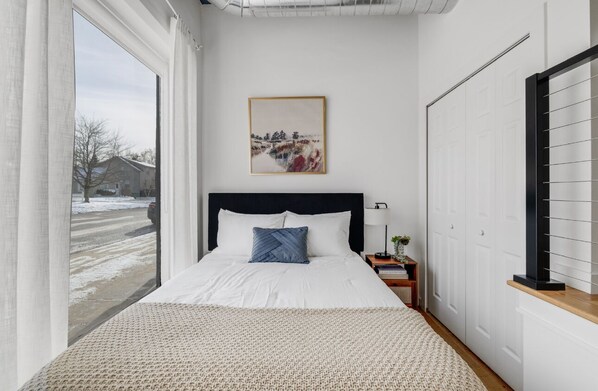 A bedroom with a large window, a bed with white linens, a blue pillow, a framed picture on the wall, and a nightstand with a lamp. - Modern Loft in Vibrant Downtown Royal Oak - Just a Block Away! (Royal Oak)