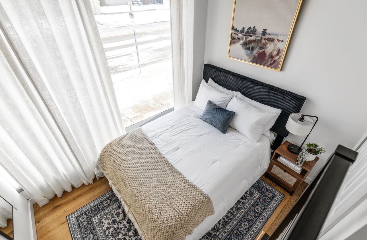 En pænt redt seng med et hvidt dynebetræk, blå og hvide puder og et beige plaide. Der er et lille natbord med en lampe og en plante. Et indrammet billede hænger over sengen.