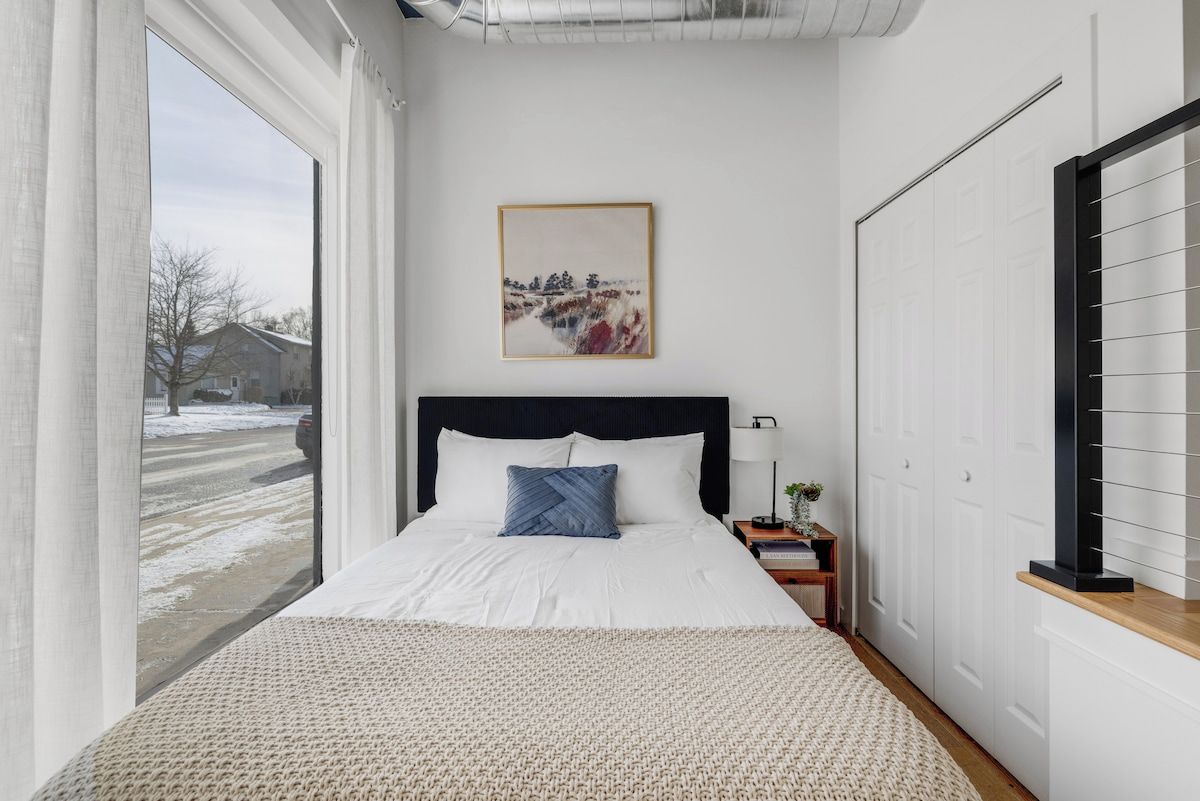 A bedroom with a large bed, a window, a door, and a painting on the wall.
