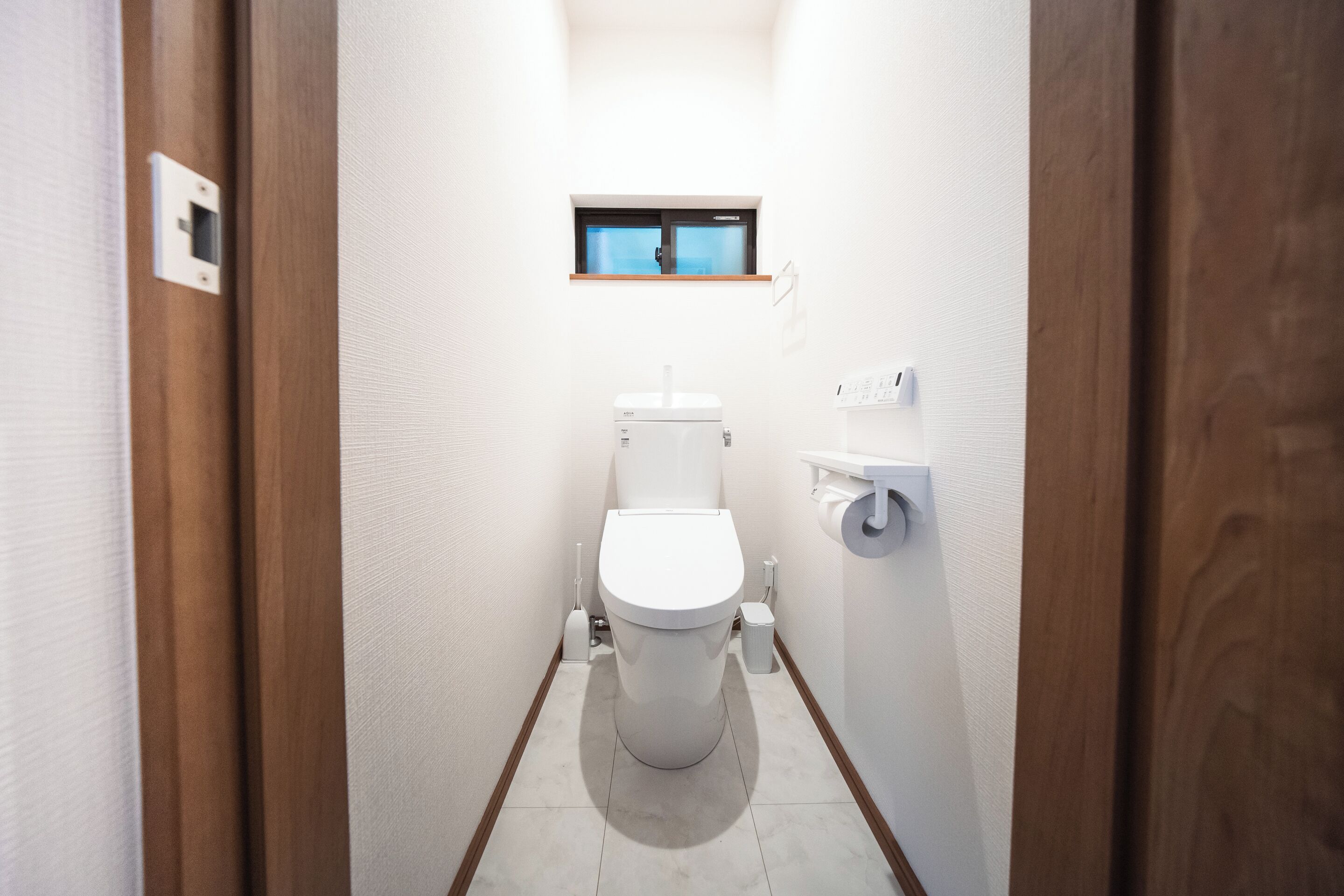 A narrow, white-walled bathroom with a toilet, a small window, and a wooden door frame.