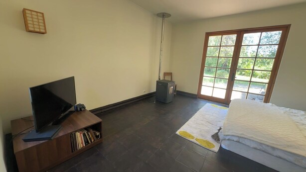 A bedroom with a TV, bookshelf, and a window with a view of greenery.