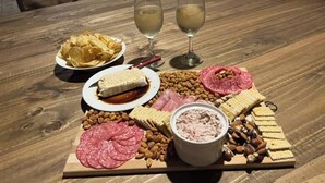 A wooden table with a charcuterie board, two glasses of white wine, and a bowl of dip.