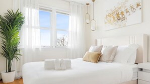 A neatly arranged bedroom with a large bed, white bedding, a bedside table, and a potted plant. - Prime Luxe 3-BR NYC - Free Parking (White Plains)