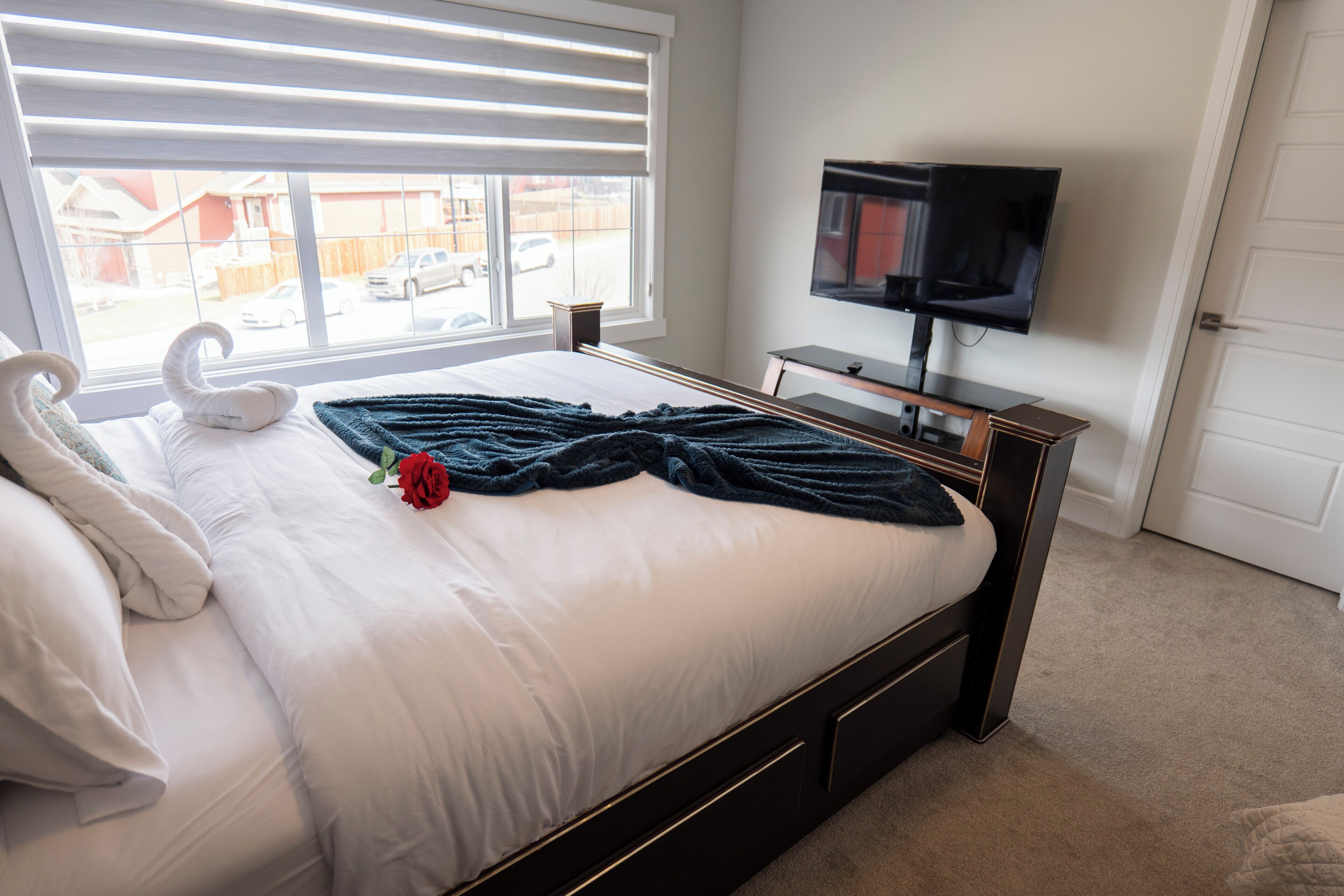 Una cama bien hecha con edredón blanco y una sola rosa roja, un televisor de pantalla plana sobre un soporte de madera, y una ventana con persianas.