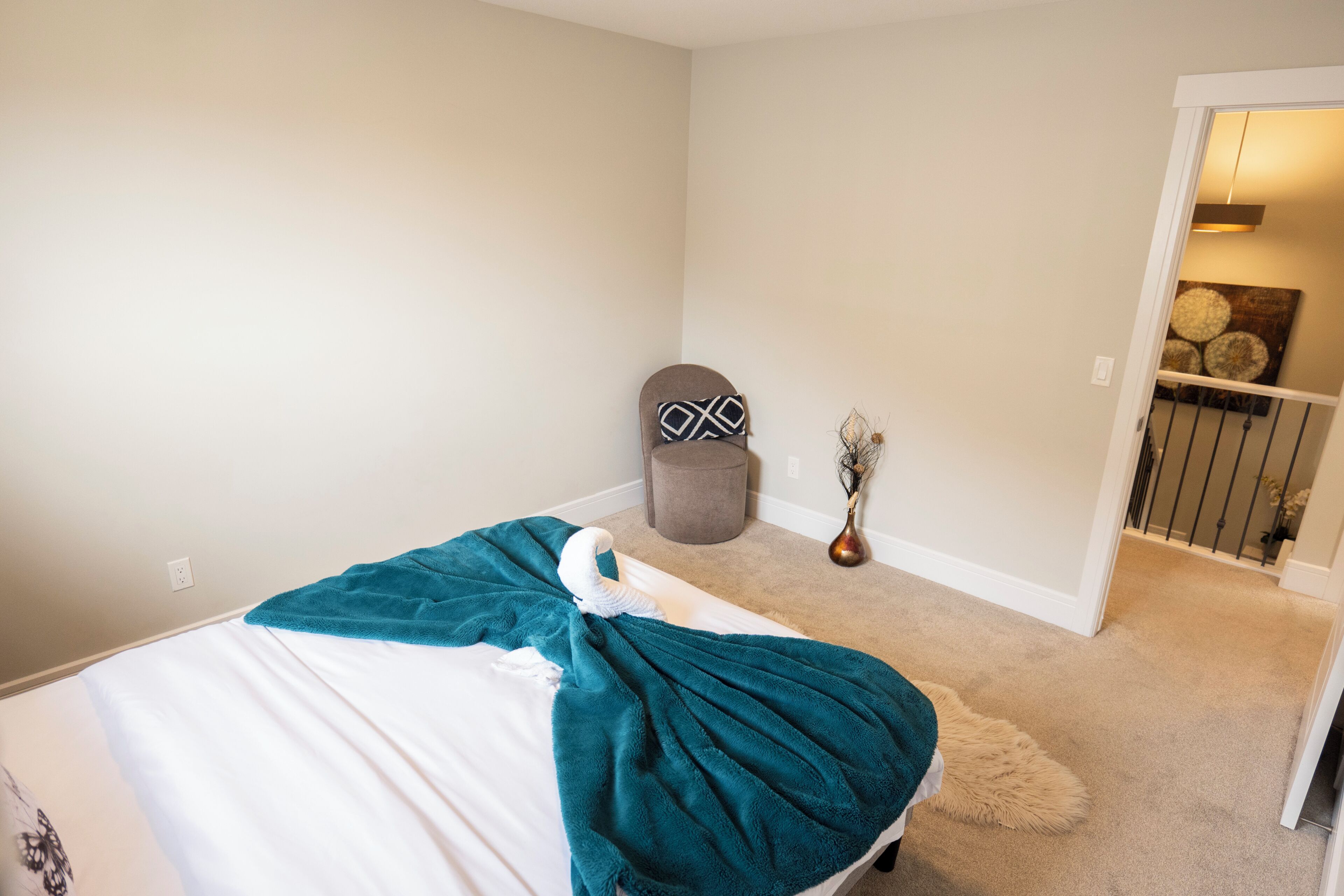 A bedroom with a bed, a grey ottoman, a vase with dried flowers, and a hallway leading to another room.