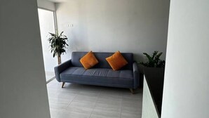 Ein modernes Wohnzimmer mit einer Couch, zwei orangen Kissen, einer Topfpflanze und einem großen Fenster.