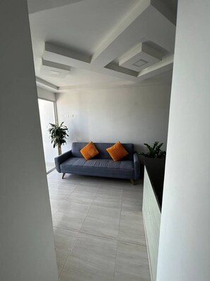 A modern living room with a sofa, two orange cushions, a potted plant, and a large window. - Alina Beautiful Cartagena Apartment (Cartagena de Indias)