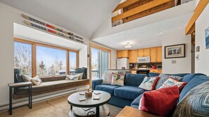 A spacious living room with a large window, a blue sofa, a wooden coffee table, and a kitchen in the background. - White Birch 58 - Ski in/out Condo, Pet friendly! (Rangeley)