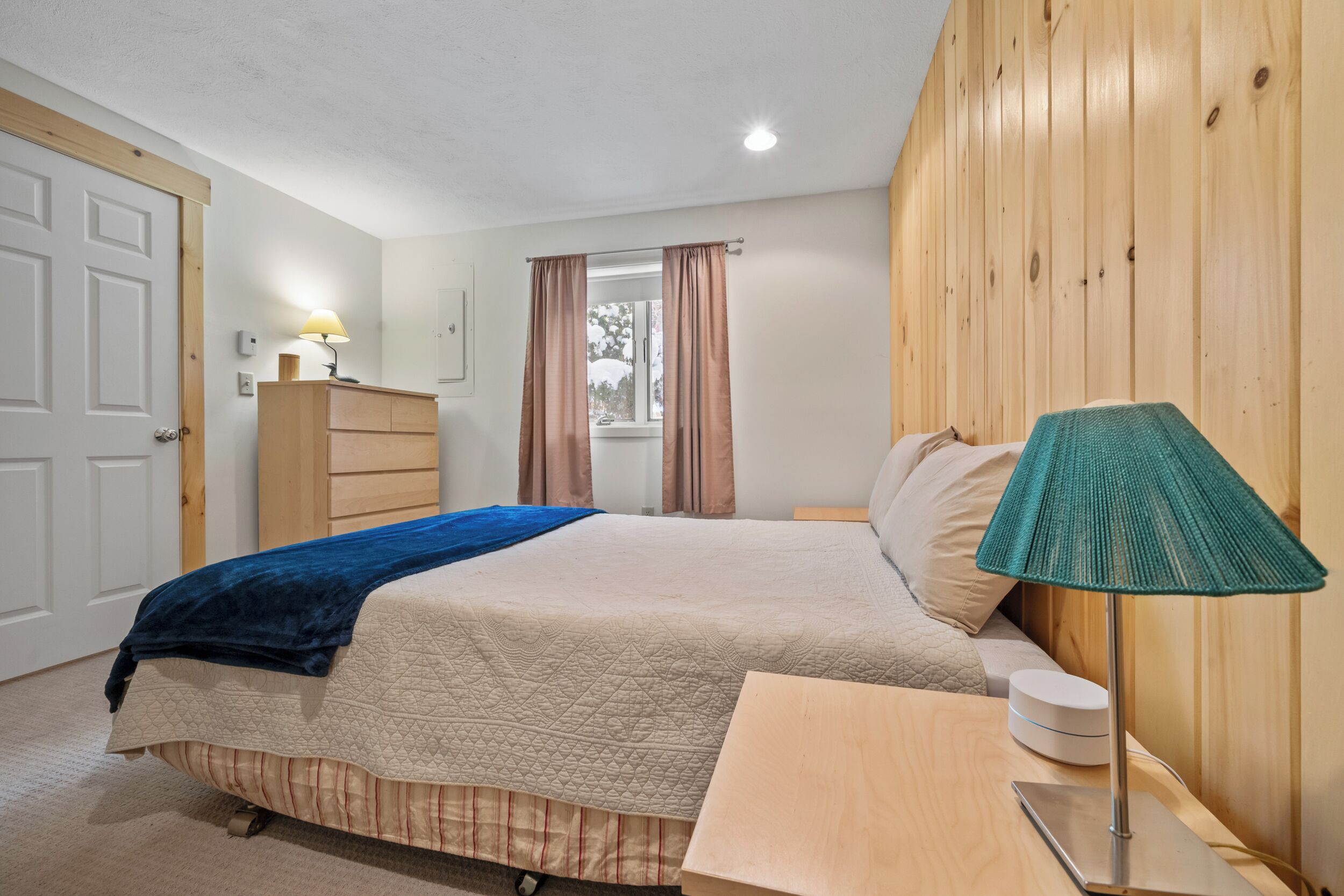 Une chambre à coucher comprenant un lit, une commode, une table de chevet, une lampe et une fenêtre avec des rideaux.