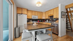 A modern kitchen with wooden cabinets, a central island with stools, stainless steel appliances, and a view of the outside. - White Birch 58 - Ski in/out Condo, Pet friendly! (Rangeley)