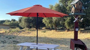 Ein Picknicktisch mit einem roten Sonnenschirm, einem Vogelhäuschen und einem hölzernen Pfosten mit einer Lampe.