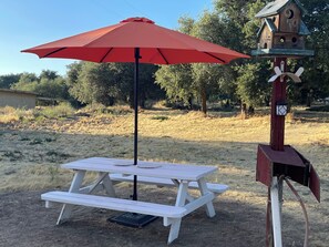 Ein Picknicktisch mit einem roten Sonnenschirm, einem Vogelhäuschen und einem hölzernen Pfosten mit einer Lampe.