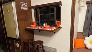Eine kleine Küche mit Fenster, Holzhockern, einem Tisch mit Unterlage und einer Futterecke für Haustiere.