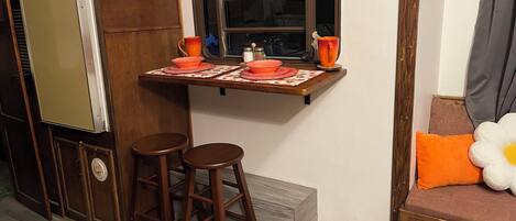 A compact kitchen with a window, wooden stools, a small table with a placemat, and a pet feeding station.