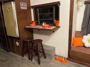 Eine kleine Küche mit Fenster, Holzhockern, einem Tisch mit Unterlage und einer Futterecke für Haustiere.