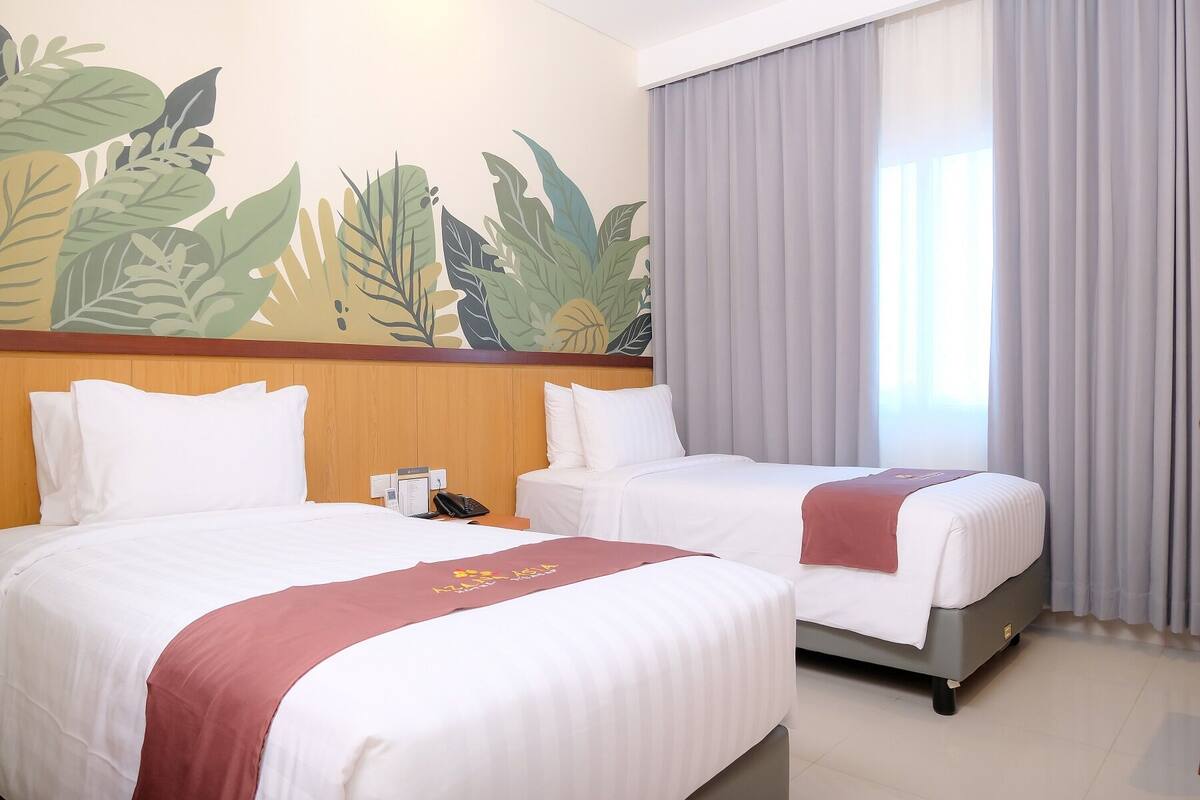 A hotel room with two beds, a wooden headboard, a tropical leaf mural, a telephone, and a window with curtains.