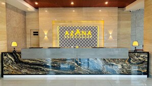 A reception desk with a marble front, a wall with 'AZANA' signage, and two lamps on either side. - Azana Asia Hotel Cilacap (Cilacap)