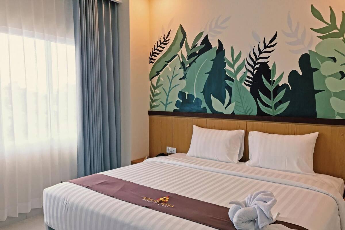 A hotel room with a bed, a wooden headboard, a wall mural of foliage, and a window with curtains.