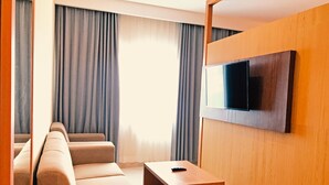 A modern hotel room with a sofa, a small wooden table, and a TV mounted on the wall. - Azana Asia Hotel Cilacap (Cilacap)