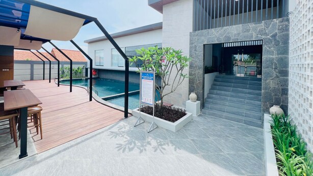 A modern outdoor pool area with a wooden deck, a covered seating area, and a stone wall with a staircase leading to an upper level. - Azana Asia Hotel Cilacap (Cilacap)