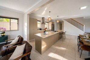A modern kitchen with a central island, a dining area with a wooden table, and a staircase leading to another level. - Family Friendly Funhouse close to All Theme Parks (Upper Coomera)