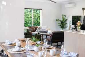 A dining area with a table set for a meal, a sofa with cushions, a ceiling fan, and a view of greenery outside. - Family Friendly Funhouse close to All Theme Parks (Upper Coomera)