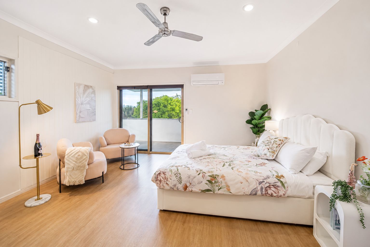 Ein Schlafzimmer mit einem Bett, einem Sessel, einem kleinen Tisch, einer Stehlampe, einem Deckenventilator und einem Fenster mit Blick ins Grüne.