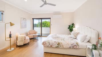 Ein Schlafzimmer mit einem Bett, einem Sessel, einem kleinen Tisch, einer Stehlampe, einem Deckenventilator und einem Fenster mit Blick ins Grüne.