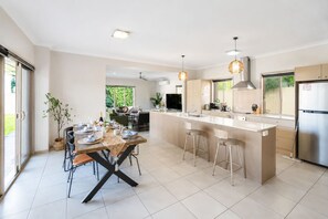 A modern kitchen with a central island, bar stools, a dining table with chairs, and a view of the outdoors. - Family Friendly Funhouse close to All Theme Parks (Upper Coomera)
