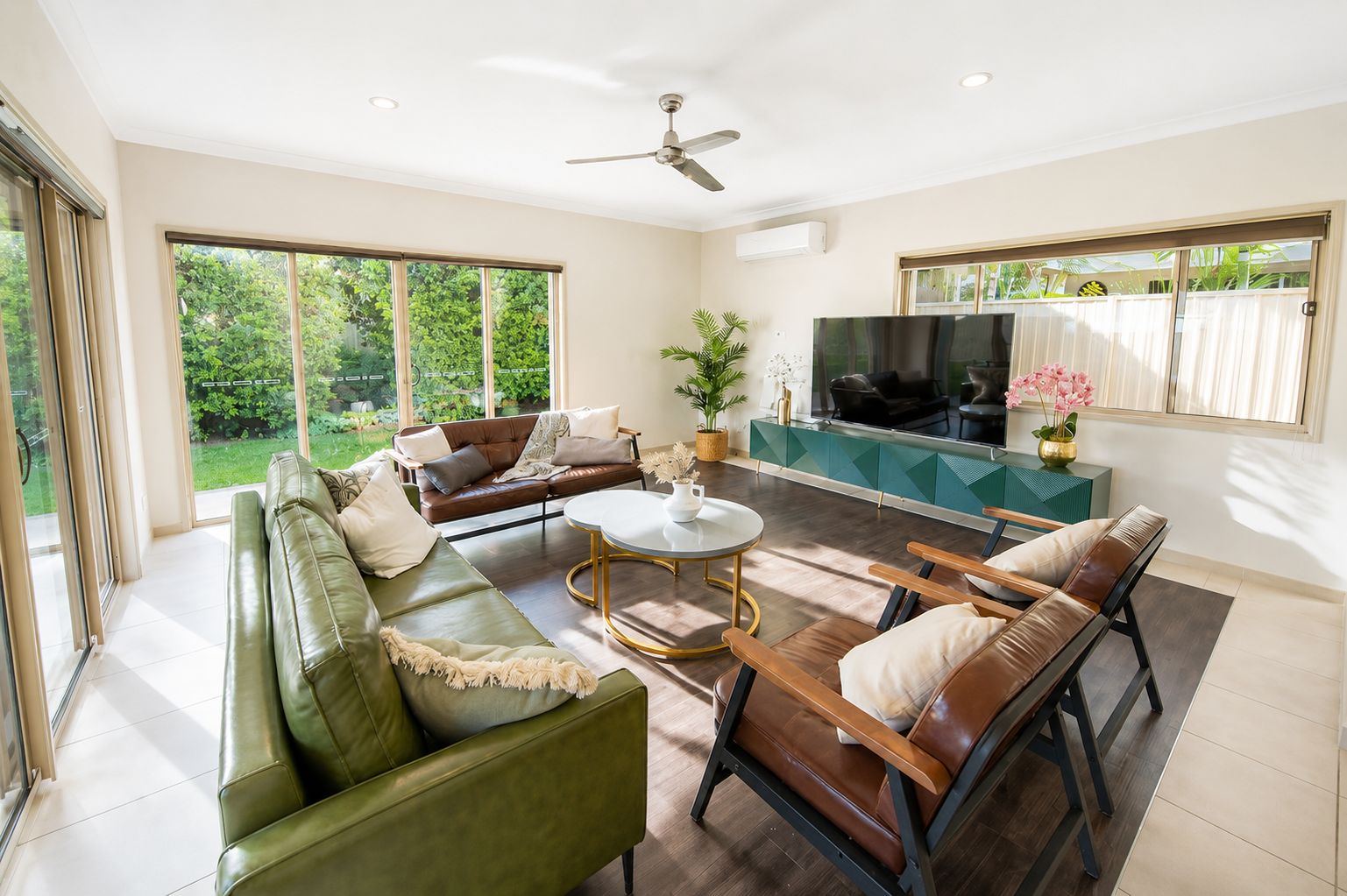 A spacious living room with a green sofa, a brown leather couch, and a wooden chair. There is a TV, a coffee table, and a potted plant.
