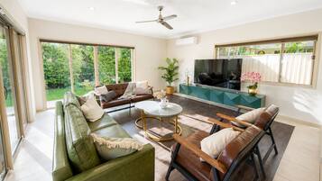 A spacious living room with a green sofa, a brown leather couch, and a wooden chair. There is a TV, a coffee table, and a potted plant.