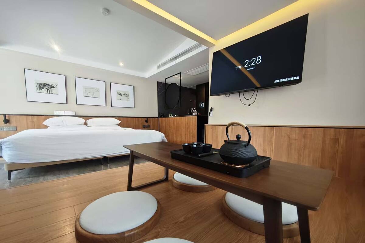 Une chambre d’hôtel moderne équipée d’un lit, d’une table avec une bouilloire, d’un téléviseur affichant l’heure et d’œuvres d’art accrochées au mur.