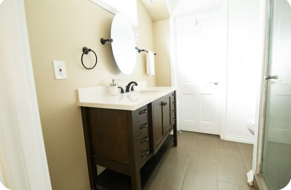 A bathroom with a dark wooden vanity, a round mirror, a white sink, and a glass shower enclosure.