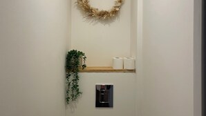 Kapea kylpyhuone, jossa on wc, seinälle kiinnitetty wc-paperiteline, pieni hylly, jossa on kaksi wc-paperirullaa, ja seinälle asennettu ilmastointilaite.