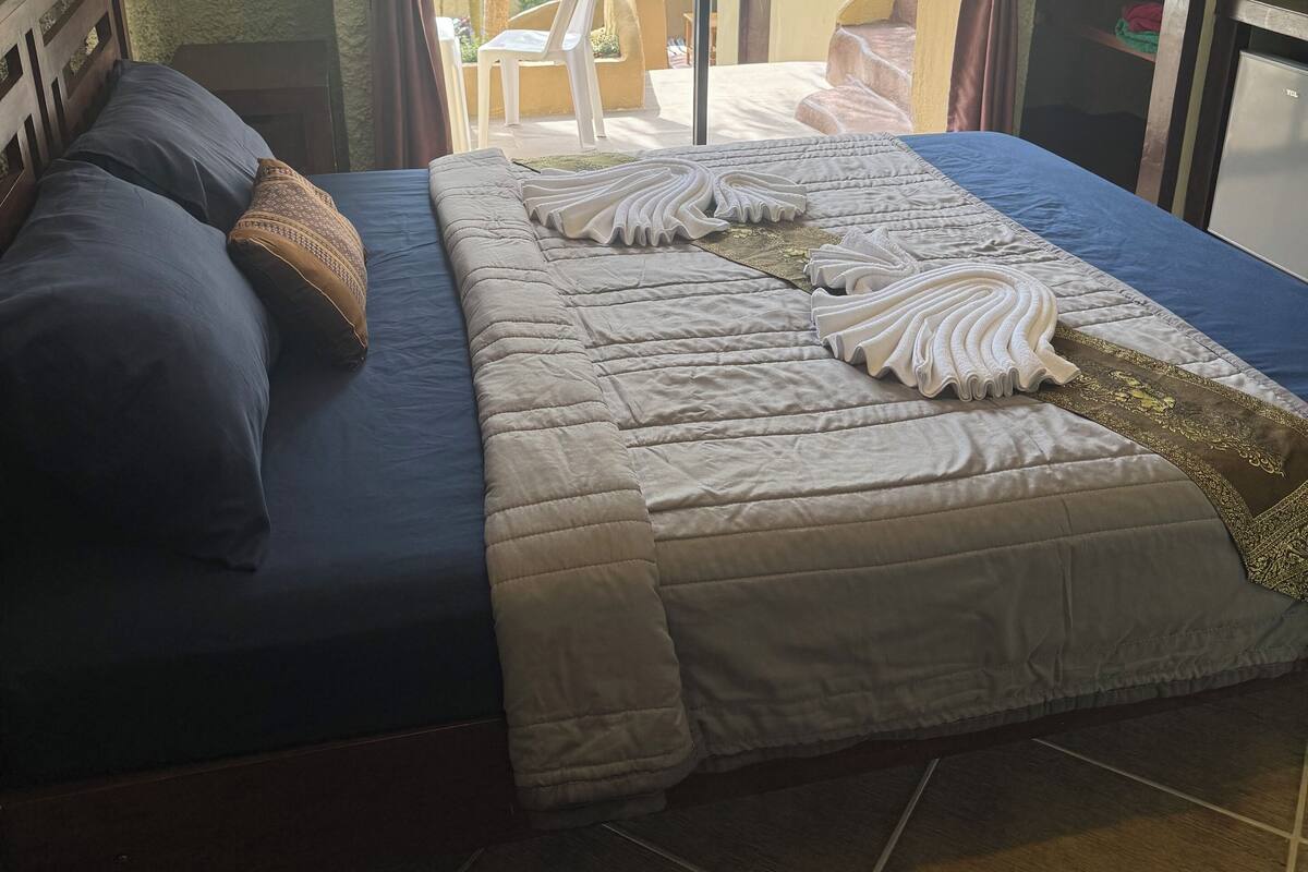 Ein Schlafzimmer mit Bett, Holz-Kopfteil, Nachttisch, Schreibtisch, Stuhl und Blick ins Freie.