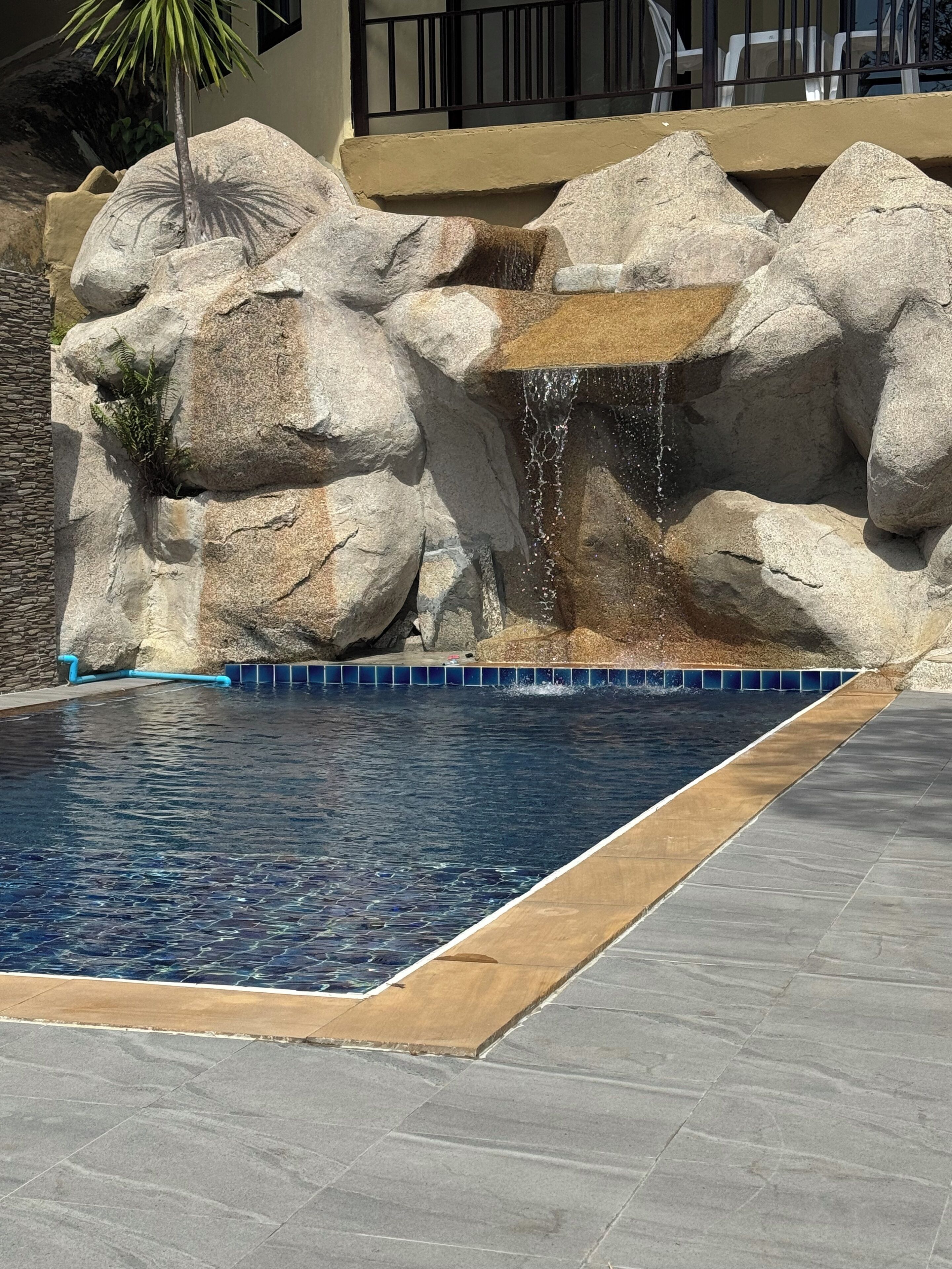 Piscina com cascata, rodeada por pedras grandes e uma borda revestida de azulejos.