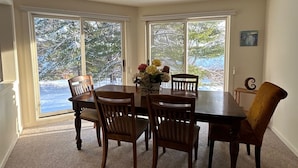 Une salle à manger avec une table ronde, six chaises en bois, un vase de fleurs et une vue sur les arbres, visible à travers de grandes portes-fenêtres coulissantes.