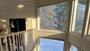 Une pièce aux hauts plafonds, dotée d’une grande fenêtre donnant sur un paysage enneigé, d’un bureau avec une chaise et d’un escalier menant à un étage supérieur.