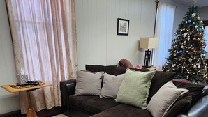 A living room with a dark brown sofa, a patterned throw blanket, a small wooden side table, and a Christmas tree. - Fountain View III- Historic Apartment Steps Away from Wellsboro Main St and Park (Wellsboro)