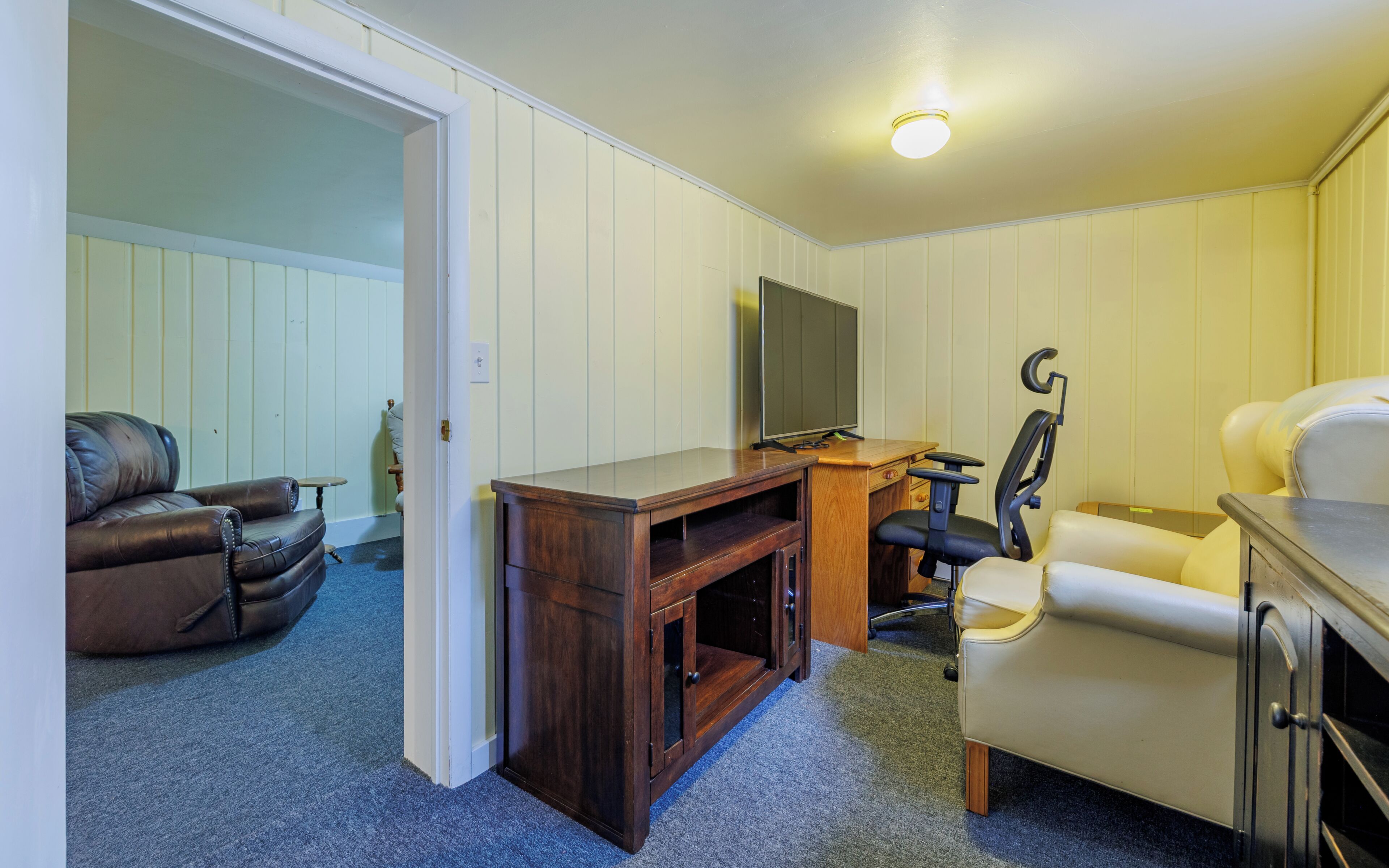 Een kamer met een bruine leren bank, een houten bureau met een computer, een zwarte bureaustoel en een televisie aan de muur.