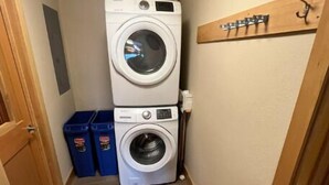 Un cuarto de lavandería con lavadoras y secadoras apiladas, un banco de madera con ganchos y contenedores azules para guardar.