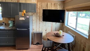 A compact kitchen with dark cabinets, a stainless steel refrigerator, a small trash bin, a round wooden table with a candle, and a window with a view of the outdoors. - Welcome to Bleistein Studio- Your launchpad for adventure (Cody)